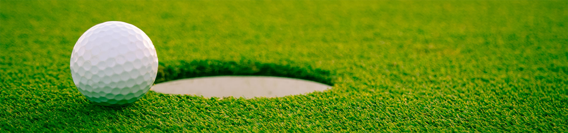 A close-up of a white golf ball resting on the green next to the edge of a golf hole on a well-manicured putting green.