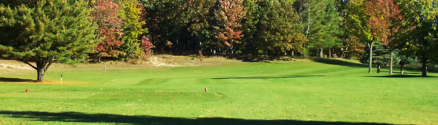 A lush, green golf course bordered by trees with autumn foliage under a clear sky; sunlight casts shadows across the grass.
